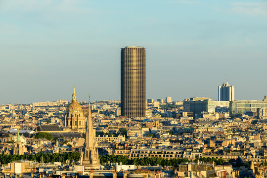 Wide Paris cityscape featuring the gleaming dome of Les Invalides and the tall Montparnasse Tower rising above layered rooftops in soft evening sunlight.