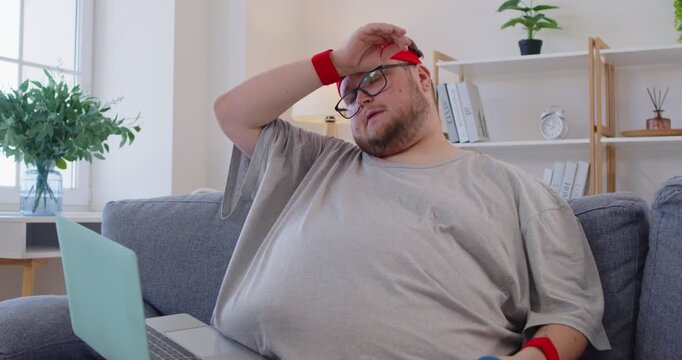 Overweight tired funny man on couch with laptop after online exercise. Fatigued, he wipes his forehead after dumbbell reps, delaying the workout at home. Lazy home fitness procrastination concept.