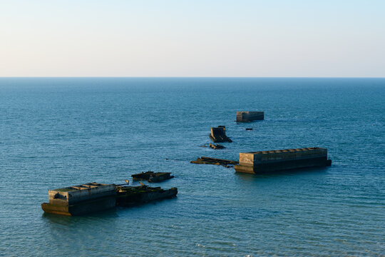 Large weathered caissons from the Mulberry Harbour rest in calm blue waters off the coast of Arromanches-les-Bains, Normandy, under soft evening light. The geometric shapes create a striking contrast