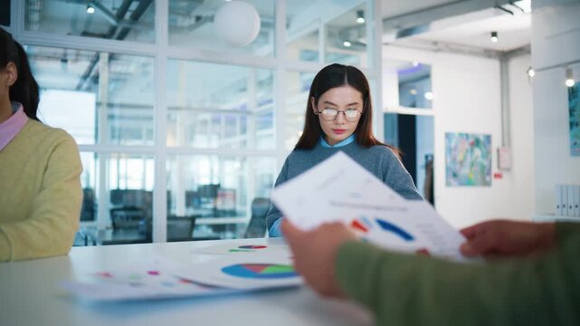 Analytical work moment at office desk. Asian woman holding tablet computer and reviewing statistics. Male colleague sorting printed charts in foreground. Bright workspace with glass walls.
