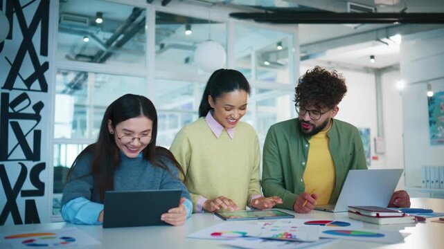 Three colleagues reviewing digital data together at desk. Pretty Caucasian woman standing in middle and checking tablet device. People discussing charts and reports in creative studio.
