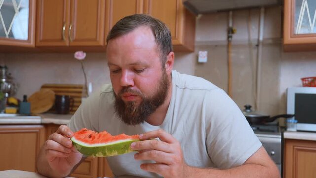 A hungry white chubby man with a beard sits at the table in the kitchen and greedily eats a large piece of ripe red watermelon, enjoying fresh and juicy summer fruits as a healthy snack