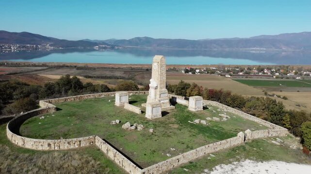 Sunny day drone footage of Doiran Memorial and Military Cemetery overlooking Lake Doiran with lion sculpture on top and rural mountain landscape beyond.