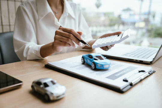 A woman reviews and signs a car insurance policy at her desk with a calculator, laptop, and miniature cars, representing auto coverage planning and financial protection.