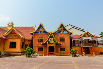 Fototapeta premium Front view of Vat Simuong temple in Vientiane, Laos, showcasing ornate gabled roofs, vibrant gold detailing, and painted Buddhist scenes. The sunlit courtyard and clear sky highlight the intricate