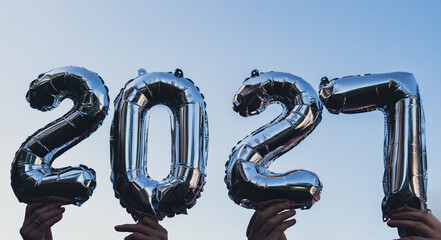Silver foil number 2027 celebration new year balloon on blue sky background. Happy New year greetings concept. Hands holding balloons two thousand twenty-fourth year Christmas holiday concept  © anna.stasiia