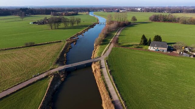 Aerial view of a rural landscape with a canal curving through green fields, two small road bridges crossing it, and a nearby house with outbuildings set among open farmland.