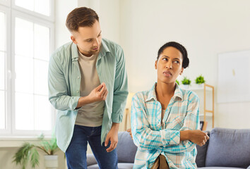 Fototapeta premium Couple conflict at home, man speaking and gesturing towards woman wife sitting on sofa, turning away head, crossing arms in annoyance, ignoring, domestic abuse, disagreement, frustration and anger