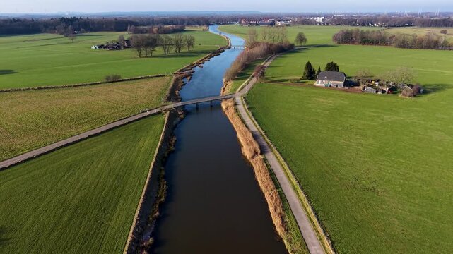 Aerial view of a rural landscape with a canal curving through green fields, two small road bridges crossing it, and a nearby house with outbuildings set among open farmland.