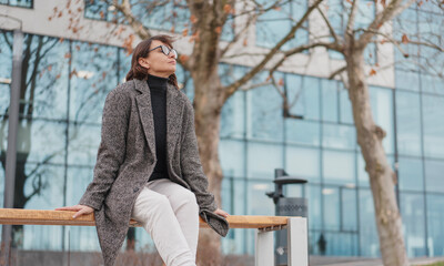 Attractive mature business woman in eyeglasses and coat resting on a bench near office building in spring