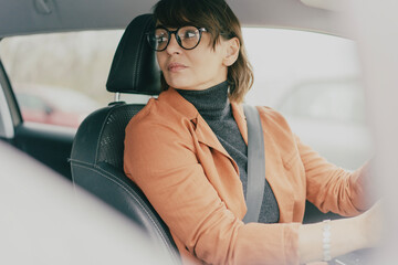 Mature business woman in blazer and eyeglasses looking back while driving or parking car