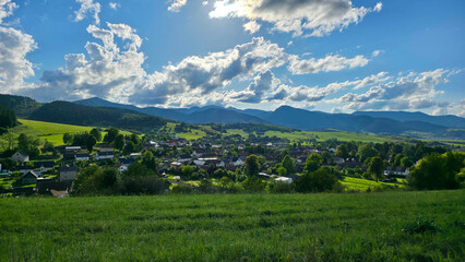Fototapeta premium Picturesque landscape view of a peaceful rural village nestled in a vibrant green valley, surrounded by rolling forested mountains under a cloudy sky.