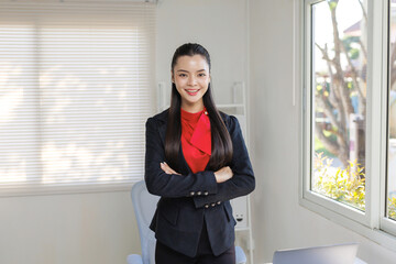 Young Asian businesswoman standing with folded arms in a bright modern office, confidently looking at the camera © amnaj