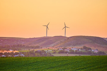 Obraz premium Wind turbines on rolling hills at sunset with farmland and orange sky