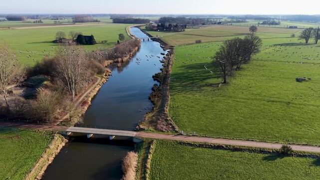 An aerial drone shot of a peaceful river winding through green fields in the Netherlands. A flock of birds flies over the water near a small bridge under a clear, bright sky.