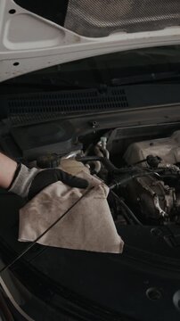 Vertical close-up of hands of anonymous male mechanic in gloves testing engine oil level with dipstick and cloth, while inspecting lubrication system during checkup in industrial workshop