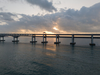 Fototapeta premium Aerial drone photo of a sea bridge at sunrise with warm sunlight, textured clouds and reflections on calm water. Scenic travel, infrastructure and cinematic background concept.