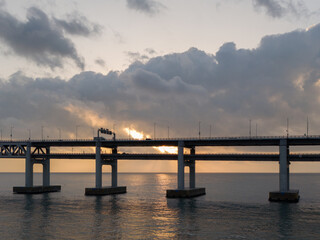 Obraz premium Aerial drone shot of a coastal sea bridge at sunrise with sunlight breaking through storm clouds and reflections on calm water. Travel, freedom, transportation and atmospheric background concept.