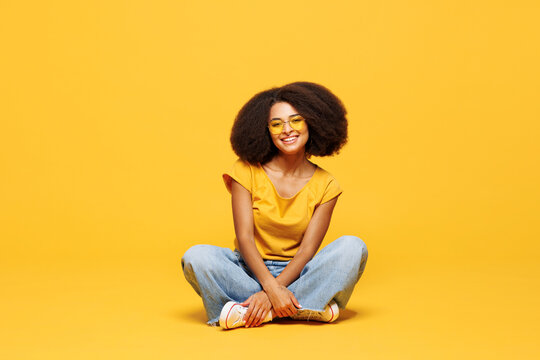 Full body young woman of African American ethnicity wear t-shirt casual clothes sunglasses sitting look camera with toothy smile isolated on plain yellow orange background studio. Lifestyle concept.