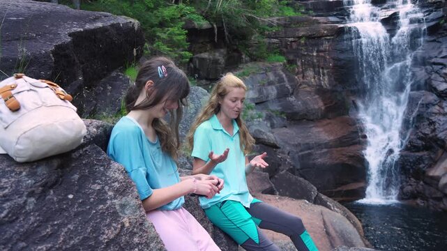 Happy mother and daughter talking by a forest waterfall in Norway. Bright northern daylight, authentic family bonding and sustainable travel concept.