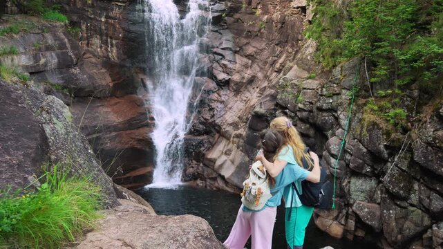 Mother and daughter admiring a misty forest waterfall in Norway from behind. Authentic back view, sustainable travel and mental health wellness through nature immersion.