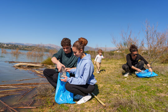 Family members picking up plastic bottles and litter from a lake with a blue sky, focused on nature conservation