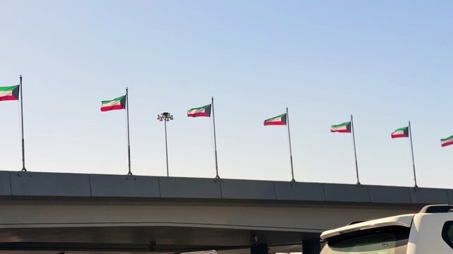Row of Kuwait national flags waving on tall flagpoles above a highway bridge under a clear blue daytime sky, symbolizing national pride and unity.