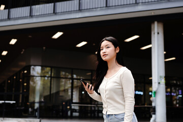Fototapeta premium asian woman college student looking around holding smartphone walking in campus