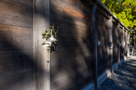 Close-up of a traditional wooden wall adorned with shide paper streamers and leafy branches at Toyouke Daijingu in Ise, illuminated by soft sunlight and surrounded by gravel and trees.