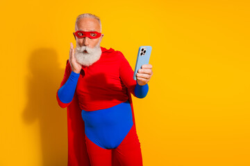 Elderly bearded man in a red and blue superhero costume takes a selfie with a blue phone against a bright yellow background at a playful event © deagreez