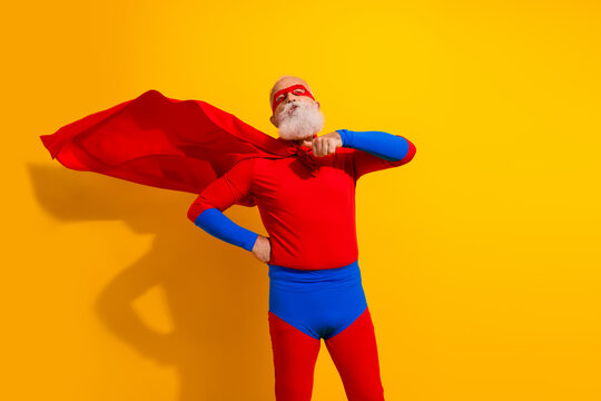 Joyful elderly man in a red and blue superhero costume poses confidently with cape against a vibrant yellow background