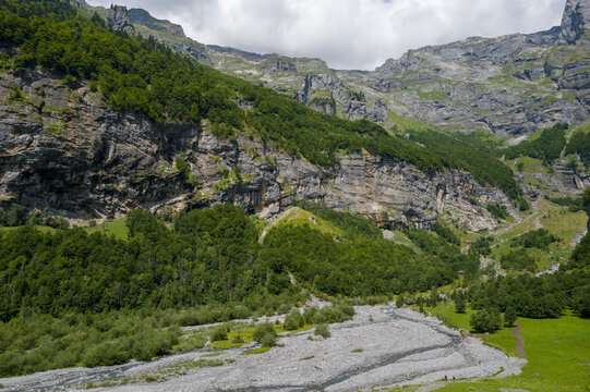 Expansive green slopes and dense forest rise to meet rugged limestone cliffs in Cirque du Fer-a-Cheval, with a stony riverbed winding through the valley under a bright summer sky.