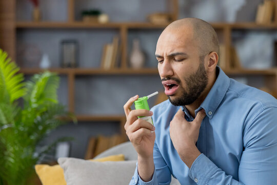 Young black man indoors applying throat spray to open mouth while touching his neck, showing discomfort from sore throat or flu as he seeks relief and self-care treatment at home