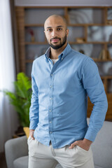 Obraz premium Bald man with a beard wearing a light blue shirt and beige pants, standing confidently with hands in pockets, looking directly at the camera in a modern home or office environment
