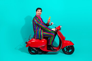 Fototapeta premium Handsome young performer on a red scooter wearing a rainbow suit smiles at the camera against a bright teal background today