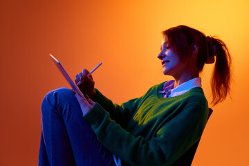 Young woman drawing on tablet with stylus in studio with vibrant gradient lighting. Concept of digital illustration work, creative freelance process and modern content production.