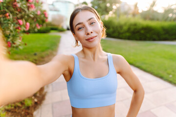 Fototapeta premium Selfie time! A happy woman takes a selfie after a workout in a sunny park. A fit woman blogs, videoconferences, and enjoys the sunny day. Concepts of sports, recreation, and blogging.