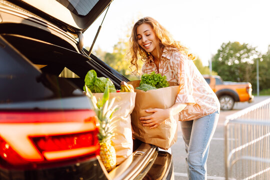 Portrait of young woman standing with fresh groceries next to her car in parking lot. Beautiful woman puts fruits, vegetables in trunk of car. Concepts of shopping, consumerism, and urban life.