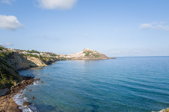 Wide angle view of Castelsardo perched atop a rocky promontory, surrounded by blue Mediterranean waters and steep coastal cliffs under a clear sky in Sardinia.