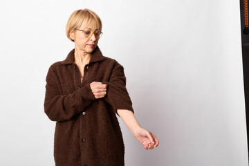 Mature woman with glasses wearing brown cozy cardigan adjusts sleeve on light background. Casual...