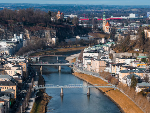 Aerial view of Salzburg, Austria, shows Salzach River with bridges, Monchsberg ridge, and Andrakirche. Winter light with bare trees and long shadows conveys a calm day.