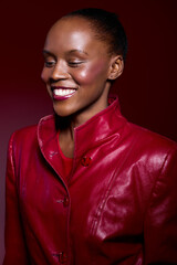 Smiling African woman with dark skin wearing a shiny red leather jacket poses against a dark red...