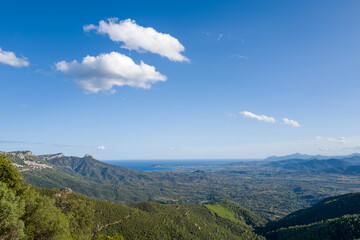 Fototapeta premium Sweeping landscape of rolling green hills stretching toward the distant blue Mediterranean Sea under a bright sky with scattered clouds near Baunei and Dorgali, Sardinia. Sunlight enhances the natural