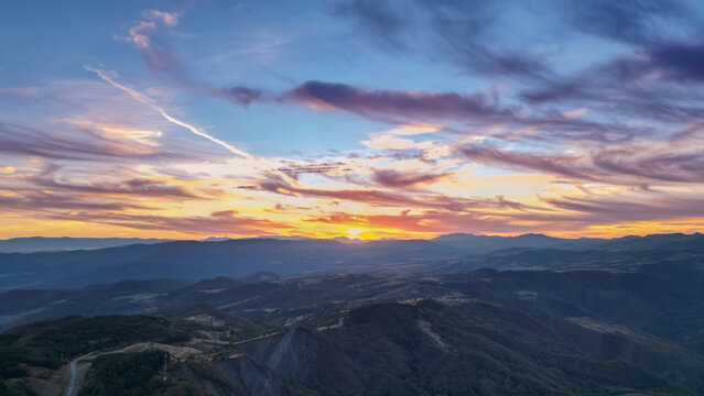 Aerial panoramic view of Georgia at sunset with golden-orange sun, colorful layered clouds, vast mountain ranges, forested slopes, and winding mountain road