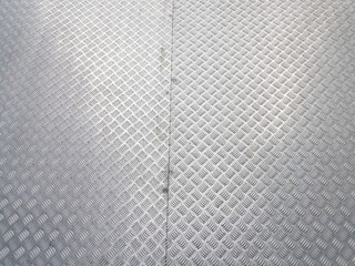 A high-angle shot of a silver aluminum checker plate floor with a repetitive five-bar diamond pattern. The metallic surface features a central vertical seam and reflects bright, natural light, creatin