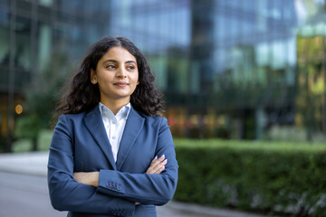 Plakat Confident young businesswoman standing outdoor with arms crossed, smiling slightly and looking away, with a modern glass office building and green foliage blurred in the background