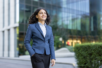 Confident young businesswoman smiling, walking outdoors with hand in pocket, against a backdrop of modern glass office buildings, representing professional success and urban career
