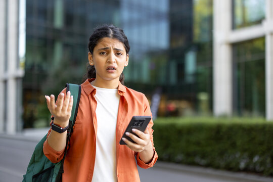 Young indian woman showing frustration and confusion while receiving bad news on her mobile phone, making a gestural expression in an outdoor urban campus setting