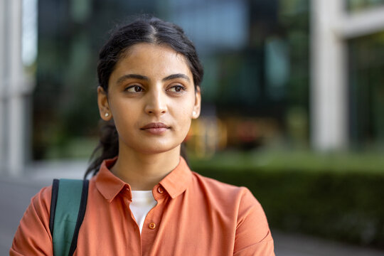 Young indian woman standing outdoors, wearing a backpack and an orange shirt, looking away with a thoughtful and serious expression, representing education, contemplation, and student life