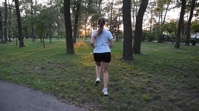 Young woman running away from camera in city park at sunset. Slow motion back view with warm evening light creates a cinematic and inspirational atmosphere. 